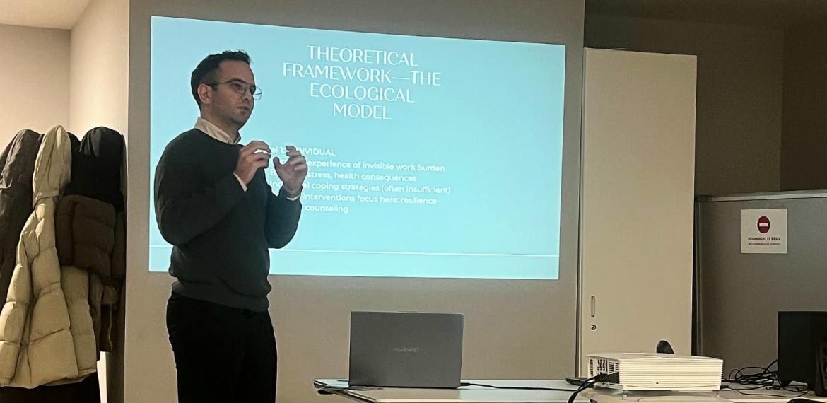 Araştırma Görevlisi Anıl Yurdakul, ayakta, sunum yaparken. önünde dizüstü bilgisayar ve arkasında projeksiyon perdesi bulunmakta. Sunumun teorik çerçevesini anlatırken çekilmiş bir fotoğraf.